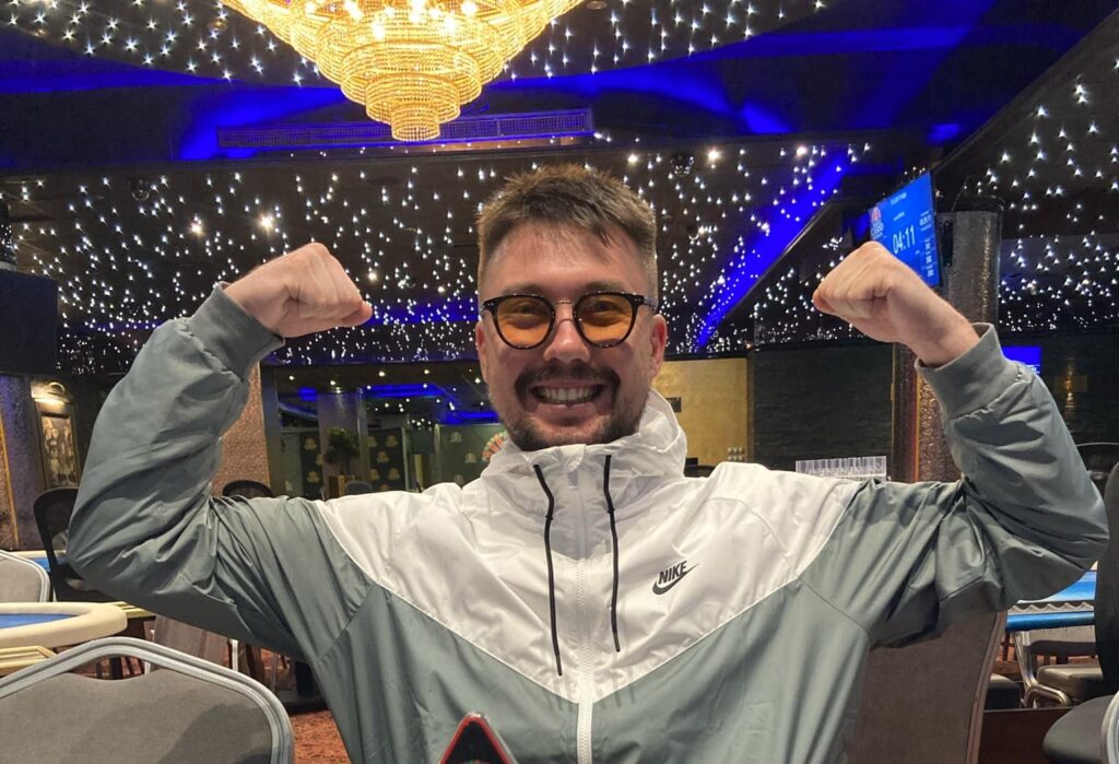 Ein Mann in einem Anzug und mit einer Sonnenbrille sitzt an einem Pokertisch und hält eine Hand mit Spielkarten. Ein Stapel Pokerchips liegt vor ihm. Im Hintergrund sind weitere Spieler und ein gr