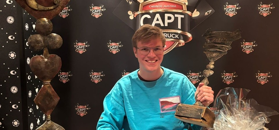 Ein Mann in einem Anzug sitzt am Pokertisch und hält stolz eine Trophäe in die Luft, umgeben von jubelnden Zuschauern.