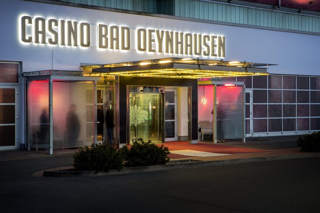 Ein Foto des Außenbereichs des Casinos Bad Oeynhausen, das von Bäumen umgeben ist und eine elegante Architektur zeigt. In der Mitte des Bildes ist der Eingang des Casinos mit großen