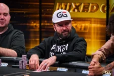 Ein Mann sitzt lächelnd an einem Pokertisch, umgeben von Spielkarten und Pokerchips. Er trägt eine schwarze Kappe und ein schwarzes Shirt. Im Hintergrund sind weitere Pokertische und Spieler