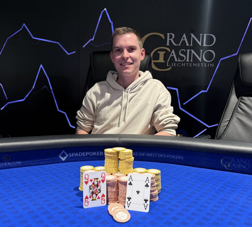 Ein eleganter Mann sitzt an einem Pokertisch und hält seine Karten in der Hand. Er trägt ein dunkles Sakko und eine Krawatte. Im Hintergrund sind Pokerchips und andere Spieler zu sehen.