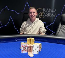 Ein eleganter Mann sitzt an einem Pokertisch und hält seine Karten in der Hand. Er trägt ein dunkles Sakko und eine Krawatte. Im Hintergrund sind Pokerchips und andere Spieler zu sehen.