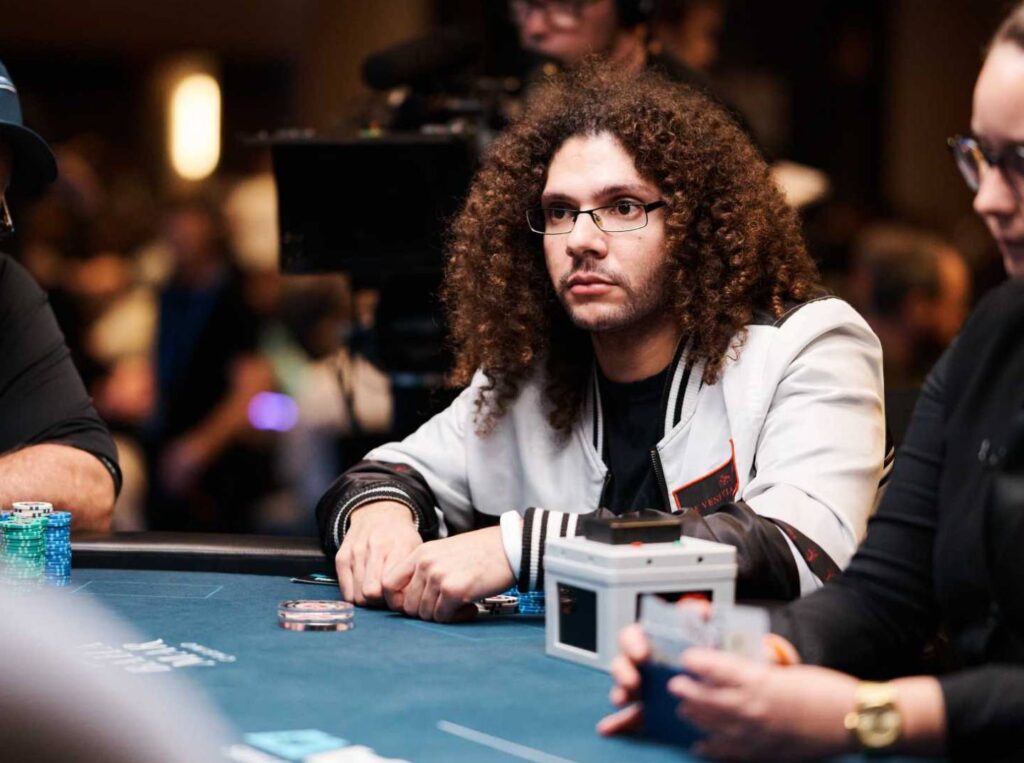 Ein Mann mit dunklen Haaren und einem blauen Hemd sitzt an einem Pokertisch und betrachtet konzentriert seine Karten. Im Hintergrund sind weitere Spieler und Chips zu sehen.