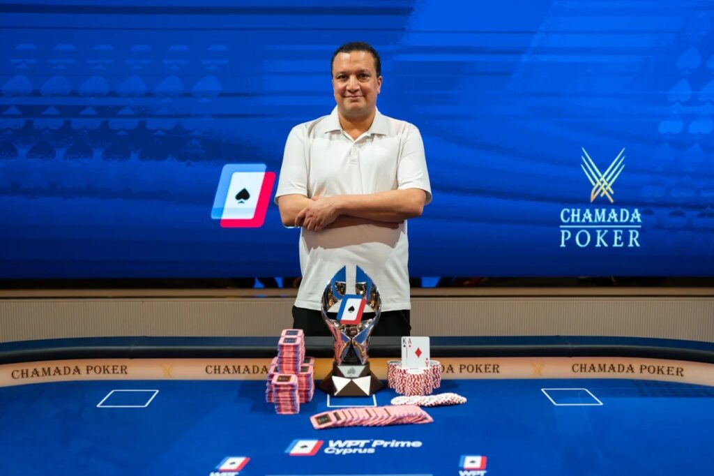 Ein Mann sitzt lächelnd an einem Pokertisch mit Chips und Karten vor sich. Er trägt ein dunkles Hemd und eine Sonnenbrille. Im Hintergrund sind andere Spieler und ein Dealer zu sehen.