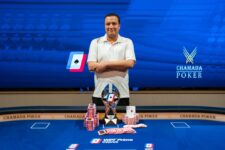 Ein Mann sitzt lächelnd an einem Pokertisch mit Chips und Karten vor sich. Er trägt ein dunkles Hemd und eine Sonnenbrille. Im Hintergrund sind andere Spieler und ein Dealer zu sehen.