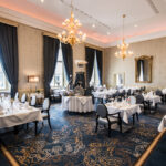 Ein elegantes Restaurant-Innenraum mit Kronleuchtern, Holztischen und Stühlen, einer Bar im Hintergrund und großen Fenstern mit Blick auf eine grüne Landschaft.