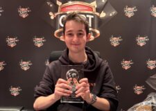 Ein Mann sitzt am Pokertisch und strahlt vor Freude, während er eine große Trophäe in die Luft hält. Um ihn herum applaudieren und gratulieren ihm andere Spieler.