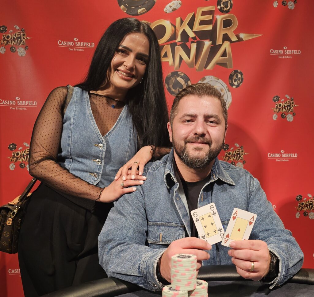 Ein Mann mit Brille und schwarzer Kappe sitzt an einem Pokertisch und hält einen großen Pokal in die Luft. Er lächelt triumphierend und hat eine Hand voller Spielchips.