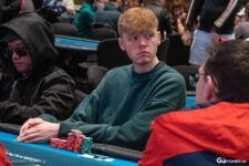 Ein Mann sitzt entspannt an einem Pokertisch und hält Karten in der Hand. Er trägt eine schwarze Brille und konzentriert sich auf das Spiel.