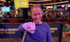 Ein Mann in einem dunklen Anzug und einer schwarzen Sonnenbrille sitzt an einem Pokertisch und hält Karten in der Hand, während er konzentriert auf sein Spiel schaut. Im Hintergrund sind weitere