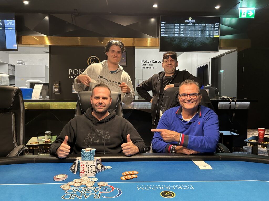 Ein Mann in einem dunkelblauen Anzug steht jubelnd mit erhobenen Armen und einem breiten Lächeln auf dem Gesicht vor einem Pokertisch. Er hält eine Hand voll Spielkarten und Chips in der Luft