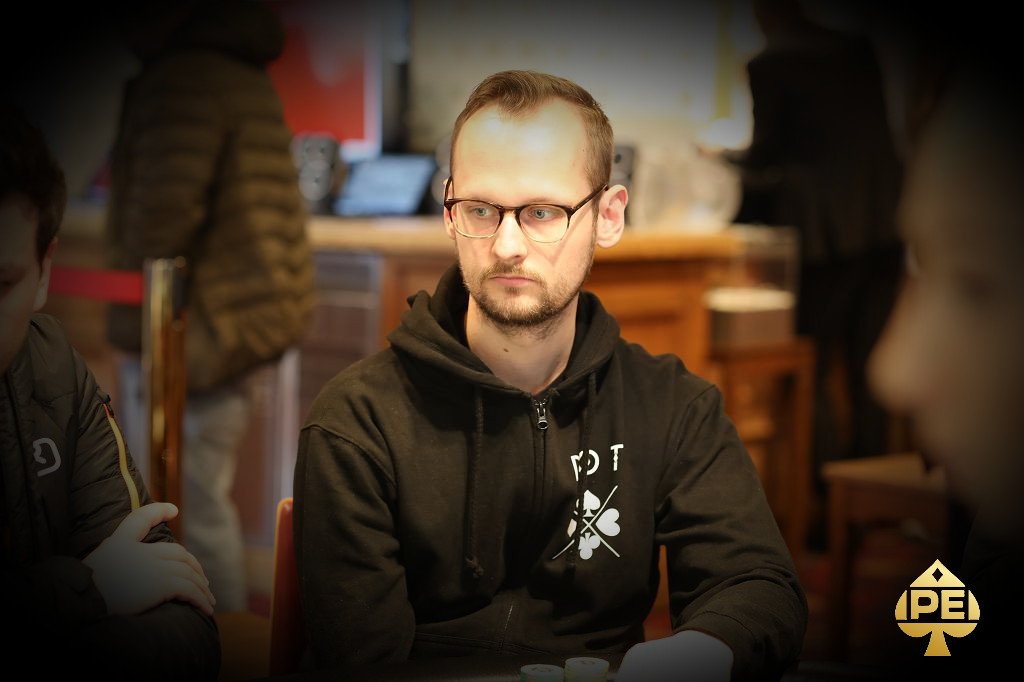 Ein Mann in einem dunklen Anzug sitzt an einem Pokertisch und hält seine Karten in der Hand, während er konzentriert auf das Spielgeschehen schaut.