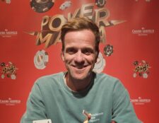 Ein Mann und eine Frau spielen Poker am Tisch in einem eleganten Casino. Der Mann hat eine Hand mit Karten in der Luft, während die Frau konzentriert ihre Karten betrachtet. Im Hintergrund sind weitere Pokert