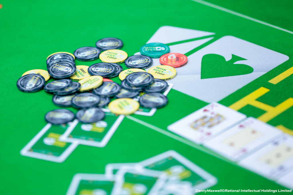 Ein Kartenspieltisch mit grüner Filzoberfläche, Pokerchips in verschiedenen Farben und Stapel von Spielkarten. Vier Personen sitzen um den Tisch herum und halten ihre Karten, während sie