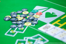Ein Kartenspieltisch mit grüner Filzoberfläche, Pokerchips in verschiedenen Farben und Stapel von Spielkarten. Vier Personen sitzen um den Tisch herum und halten ihre Karten, während sie