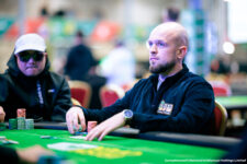 Ein Mann spielt Poker an einem Tisch in einem Casino, umgeben von anderen Spielern. Er hat eine ernste Miene und hält Karten in der Hand, während er auf seine Gegner blickt. Das Bild zeigt eine