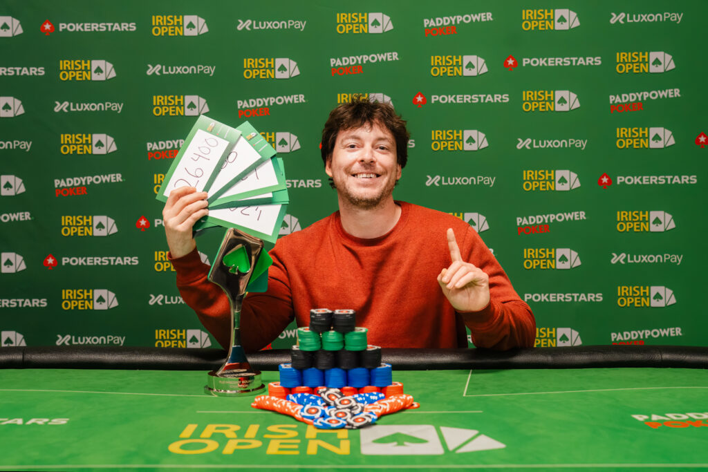 Ein Mann sitzt an einem Pokertisch und hält seine Karten in der Hand. Er hat eine schwarze Kappe auf und trägt ein schwarzes Oberteil. Im Hintergrund sind weitere Pokertische und Spieler zu sehen