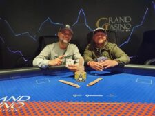 Ein Mann sitzt an einem Pokertisch und hält eine Hand mit Spielkarten. Er hat einen ernsten Blick und trägt einen Anzug. Neben ihm liegen Pokerchips und ein Getränk. Im Hinter