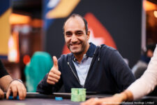 Ein Mann in einem dunkelblauen Anzug sitzt an einem Pokertisch und schaut konzentriert auf seine Karten. Er hat dunkles Haar und trägt eine Uhr am Handgelenk. Im Hintergrund