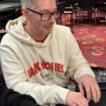 Ein professioneller Pokertisch mit grünen Filz und einem Stapel aus Pokerchips in verschiedenen Farben, umgeben von Spielkarten und Getränken. Zwei Spieler sitzen mit ernstem Gesichtsausdr