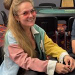 Ein Mann und eine Frau spielen Poker in einem eleganten Casino. Der Mann hat einen Anzug an und die Frau trägt ein rotes Abendkleid. Der Tisch ist mit Pokerchips und Karten bedeckt. Im H