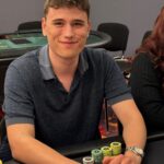 Ein Spieler hält zwei Spielkarten in der Hand während er an einem Pokertisch sitzt, umgeben von Pokerchips und anderen Spielern.