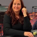 Ein professioneller Pokerspieler mit Sonnenbrille und Kappe, der konzentriert an einem Pokertisch sitzt und seine Karten betrachtet. Im Hintergrund sind weitere Spieler und Pokerchips zu sehen.