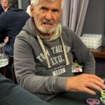 Ein Mann und eine Frau sitzen an einem Pokertisch und spielen Karten. Der Mann hat eine Hand mit Chips und schaut konzentriert auf seine Karten, während die Frau mit einem selbstbewussten Lächeln ihre K
