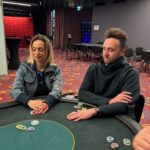 Ein Bild eines Pokertisches mit Spielkarten, Pokerchips und Getränken, umgeben von Menschen, die Poker spielen und Spaß haben.