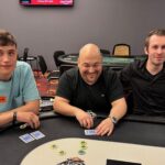 Ein professioneller Pokerspieler mit Sonnenbrille und Kappe sitzt an einem Pokertisch und hält seine Karten in der Hand, während er konzentriert auf seine Gegner schaut.