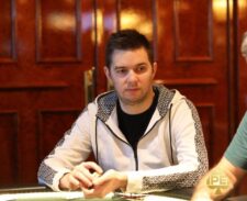 Ein Mann mit dunklen Haaren und einem schwarzen T-Shirt sitzt an einem Pokertisch und betrachtet konzentriert seine Karten. Im Hintergrund sind weitere Spieler und Karten auf dem Tisch zu sehen.