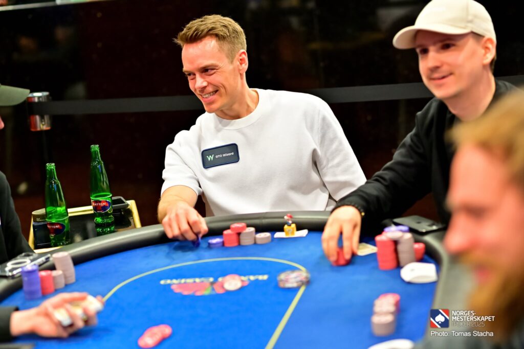 Ein junger Mann sitzt an einem Pokertisch und betrachtet konzentriert seine Karten. Er hat blonde Haare und trägt ein blaues Hemd. Im Hintergrund sind weitere Pokerspieler zu sehen.