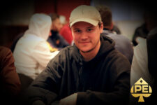 Ein Mann sitzt an einem Pokertisch und hält Karten in der Hand. Er trägt einen dunklen Anzug und hat kurze, dunkle Haare.