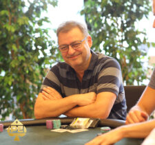 Ein Mann mit dunklen Haaren und einem schwarzen Anzug sitzt an einem Pokertisch und hat einen ernsten Gesichtsausdruck. Er hält Karten in der Hand und schaut konzentriert auf den T
