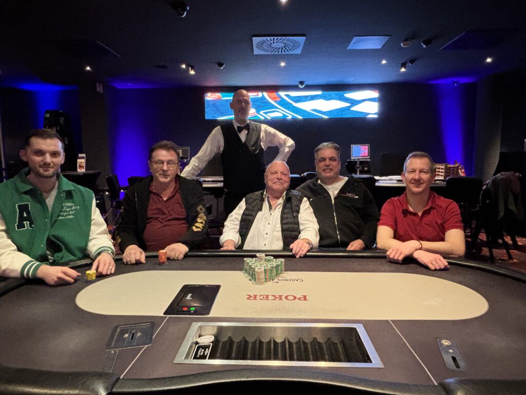 Ein Mann in einem schwarzen Anzug spielt Poker an einem eleganten Tisch mit grünem Filz und hält dabei seine Spielkarten in der Hand. Im Hintergrund sind mehrere Spieler und eine Glühbirne zu