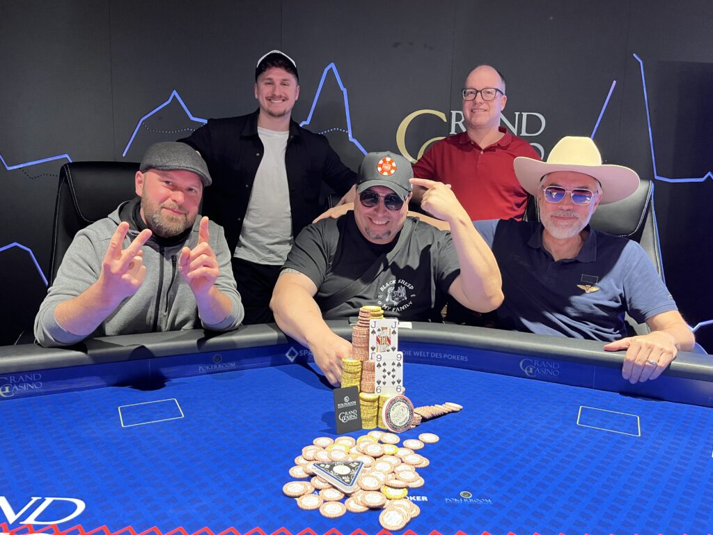 Ein Mann und eine Frau sitzen an einem Pokertisch und spielen ein intensives Spiel. Der Mann hat eine Hand mit Karten in der Luft, während die Frau konzentriert ihre Karten betrachtet. Im Hintergrund sind