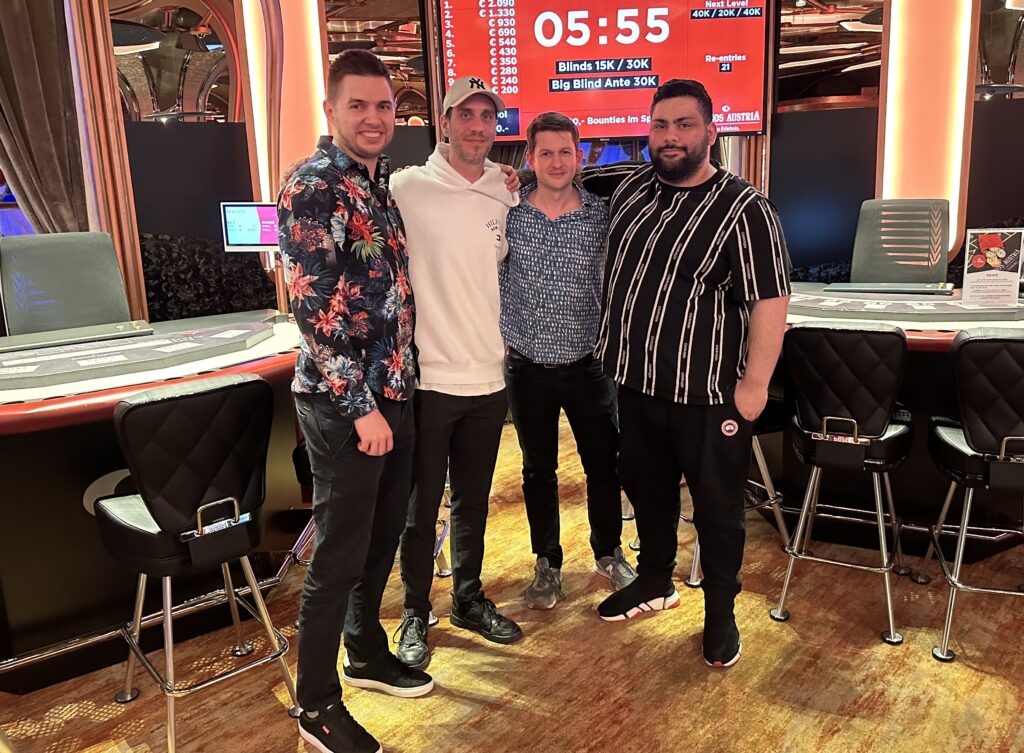 Ein Mann sitzt an einem Pokertisch und hält zwei Spielkarten in der Hand. Er schaut konzentriert auf den Tisch, umgeben von Pokerchips und anderen Spielern im Hintergrund.