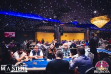 Ein professioneller Pokerspieler sitzt an einem Tisch während eines Turniers und betrachtet konzentriert seine Karten. Um ihn herum sind weitere Spieler und Zuschauer zu sehen. Die Spannung ist förmlich