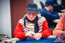 Ein Mann mit dunklen Haaren und einem schwarzen Anzug sitzt entspannt an einem Pokertisch und schaut nachdenklich auf seine Karten. Er hat eine ernste Miene und wirkt konzentriert.