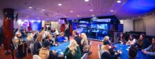 Ein Mann und eine Frau sitzen an einem Tisch, der mit Pokerchips und Spielkarten bedeckt ist. Sie schauen konzentriert auf einen großen Bildschirm, auf dem ein Pokerturnier live gestreamt