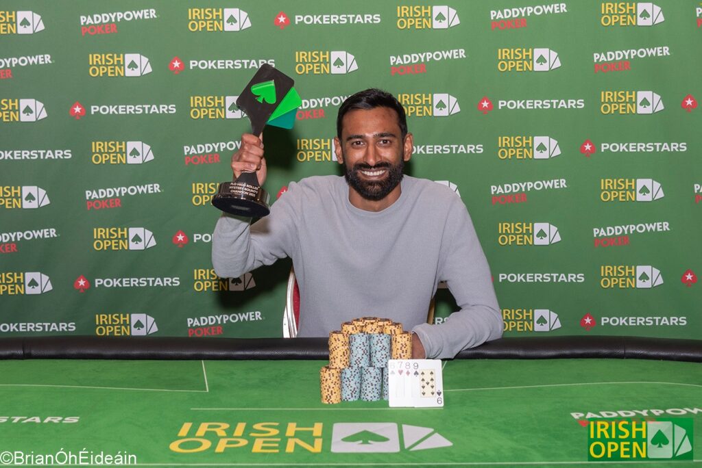 Ein Mann mit dunklem Haar und Bart, gekleidet in einem schwarzen T-Shirt und einer Kappe, posiert mit einem Pokal in der Hand vor einem Hintergrund mit Pokertischen und anderen Spielern.