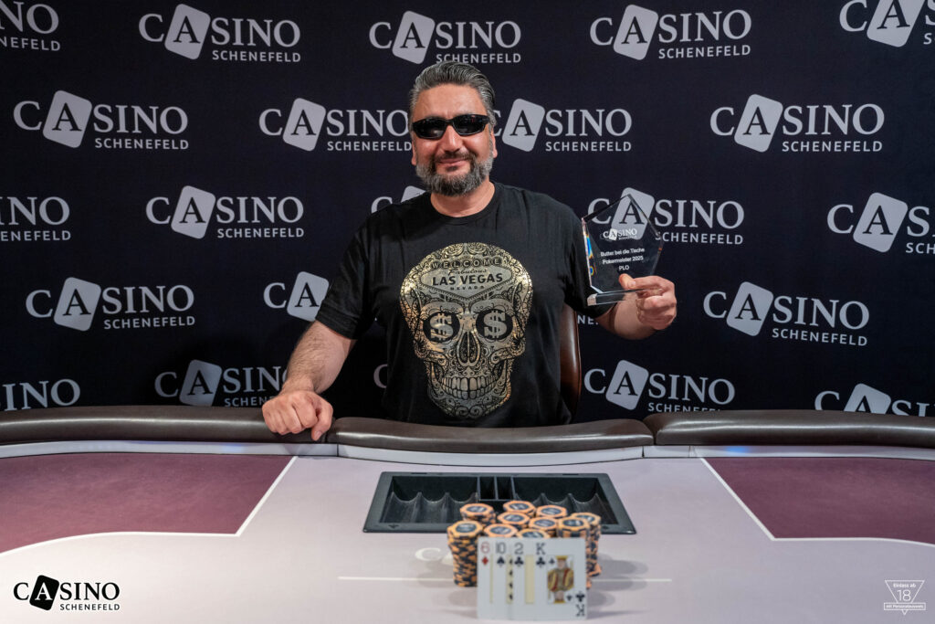 Ein Mann mit einem breiten Lächeln und einer Sonnenbrille sitzt an einem Pokertisch, umgeben von Chips und Spielkarten. Er hält triumphierend eine Gewinntrophäe in die Luft.