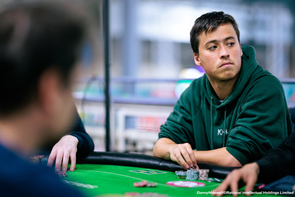 Ein Mann mit dunklem Haar und Bart sitzt an einem Pokertisch und hält Karten in der Hand. Er trägt ein dunkles Hemd und eine schwarze Baseballkappe. Im Hintergrund sind andere Pokerspi