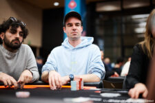 Ein Mann in einem schwarzen Anzug und einer schwarzen Sonnenbrille sitzt lässig an einem Pokertisch in einem Casino. Er hält Karten in der Hand und schaut konzentriert auf seine Gegner.