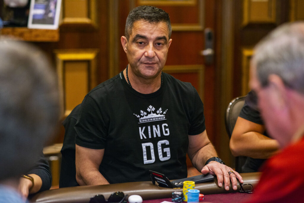 Ein Mann sitzt lächelnd an einem Pokertisch, umgeben von Chips und Karten. Er trägt Kopfhörer und eine schwarze Jacke.