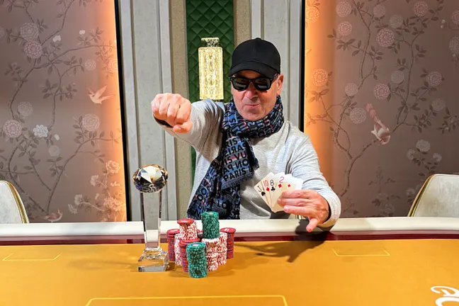 Ein Mann in einem dunklen Anzug und einer roten Krawatte sitzt an einem Pokertisch und hält eine Hand mit Spielkarten. Er schaut konzentriert auf seine Karten. Im Hintergrund sind andere