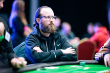 Ein junger Mann in einem schwarzen Anzug sitzt an einem Pokertisch mit Chips und Karten vor sich. Er schaut konzentriert auf seine Karten.