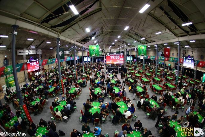 Ein Mann sitzt an einem Pokertisch bei einem Turnier. Er trägt eine grüne Jacke und hat ein Dummy vor sich liegen. In der Hand hält er Spielkarten und schaut konzentriert auf seine