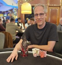 Ein Mann in einem dunkelblauen Anzug und einer roten Krawatte sitzt an einem Pokertisch und hält zwei Spielkarten in der Hand. Im Hintergrund sind Pokerchips und andere Spieler zu sehen.