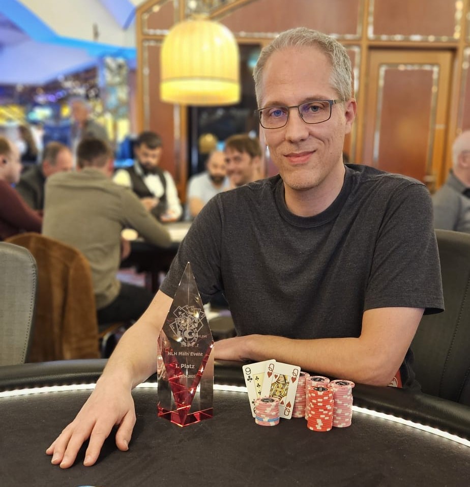 Ein Mann in einem dunkelblauen Anzug und einer roten Krawatte sitzt an einem Pokertisch und hält zwei Spielkarten in der Hand. Im Hintergrund sind Pokerchips und andere Spieler zu sehen.